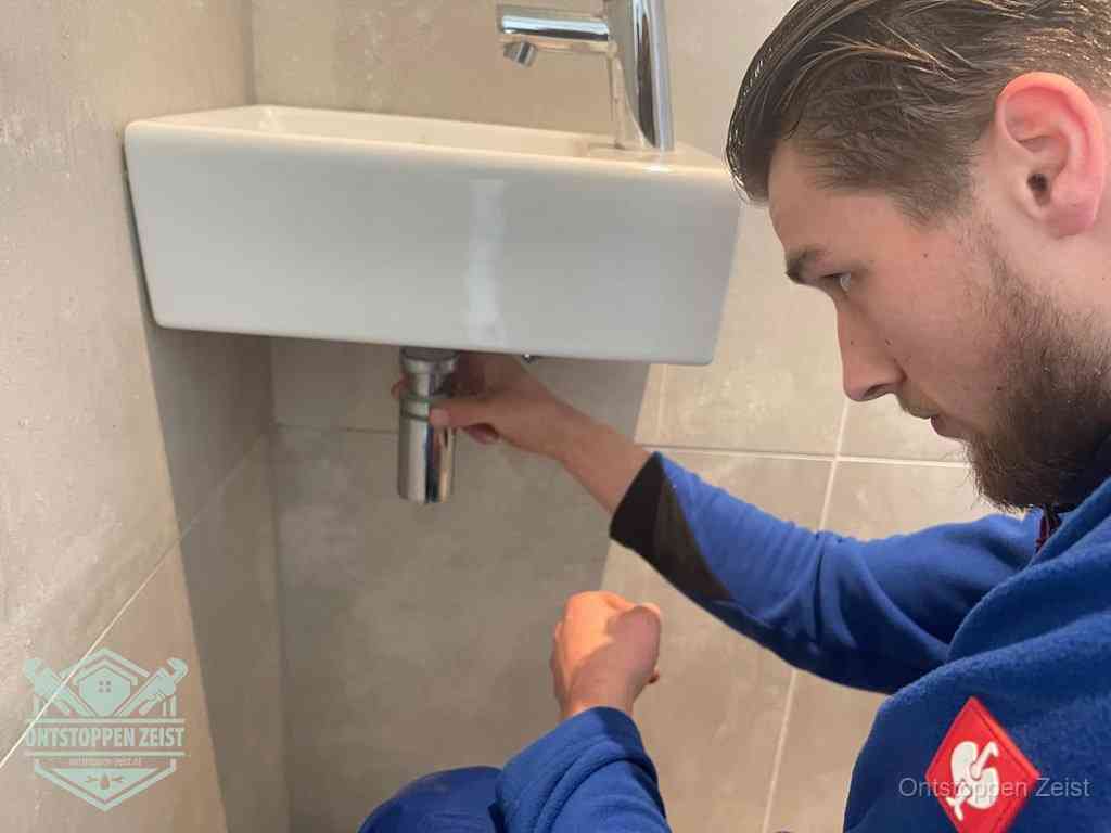 Een monteur draait met de hand de zwanenhalssifon onder een fonteinwasbak in de toiletruimte los om de bron van rioollucht in Zeist te vinden Een monteur draait met de hand de zwanenhalssifon onder een fonteinwasbak in de toiletruimte los om de bron van rioollucht in Zeist te vinden